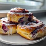 Three decadent blueberry cheesecake rolls stacked on a white plate, drizzled with white icing.