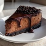 A rich slice of chocolate cheesecake with a dark chocolate crust, topped with glossy ganache and cookie crumbs.