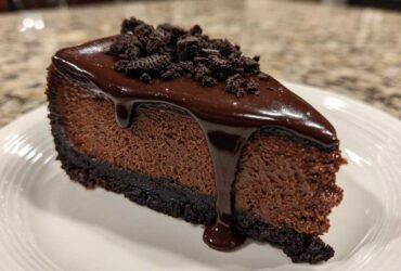 A rich slice of chocolate cheesecake topped with glossy chocolate ganache and cookie crumbs.