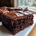 A close-up, angled shot of a rich, dark chocolate slice of Decadent Frosted Zucchini Brownies on a white plate.