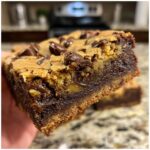 A close-up view of a thick, chewy bar showing the distinct layers of a decadent fudgy brookie, topped with chocolate chips.