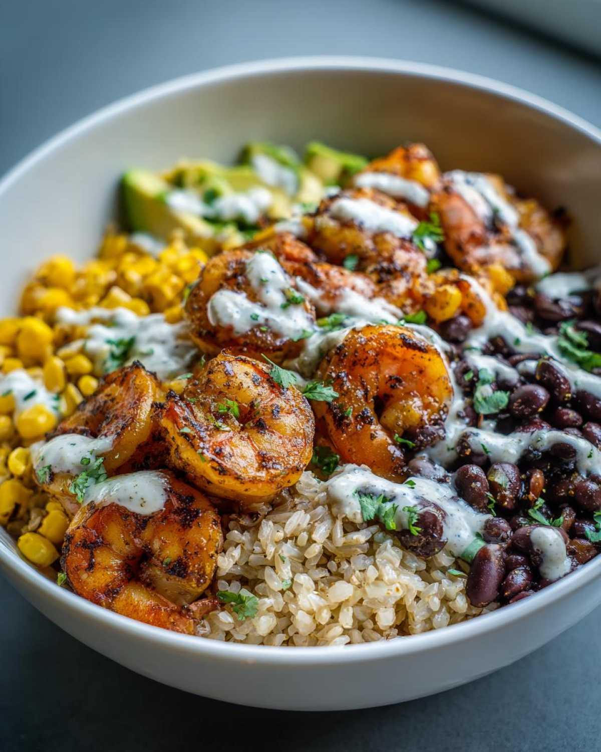 Close-up of a delicious shrimp bowl featuring seasoned shrimp, brown rice, black beans, corn, avocado, and a white sauce drizzle.