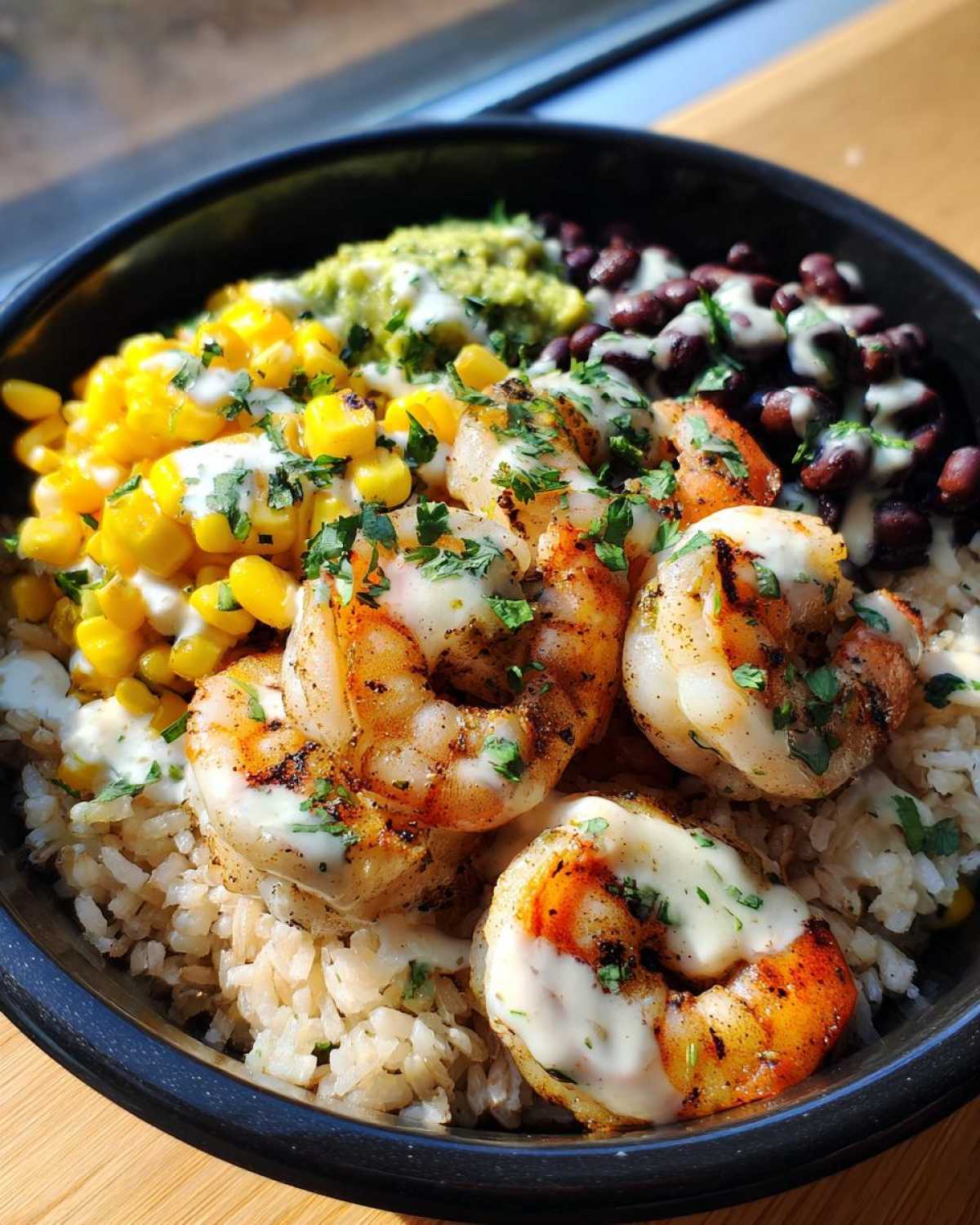 Close-up of a vibrant shrimp bowl featuring grilled shrimp over brown rice, topped with corn, black beans, guacamole, and a creamy sauce.