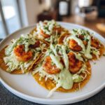 Four assembled shrimp taco servings on corn tortillas topped with cabbage slaw, seasoned shrimp, and creamy green sauce.