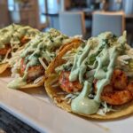 Three assembled shrimp taco servings topped with shredded cabbage and a generous drizzle of creamy green avocado sauce.