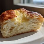 A close-up of a thick slice of fluffy beer bread, glistening with melted butter on top, sitting on a white plate.