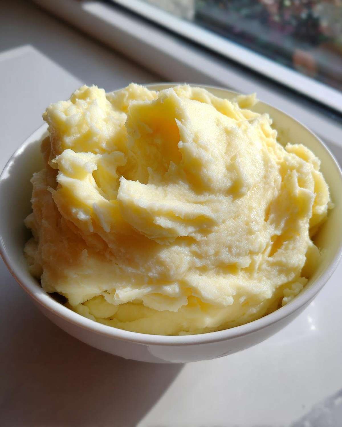 A close-up of a white bowl filled with fluffy, creamy mashed potatoes, brightly lit by a window.