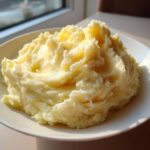 A generous serving of fluffy, creamy mashed potatoes piled high on a white plate, catching bright sunlight.
