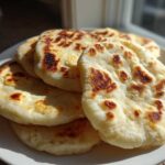 A stack of freshly cooked, slightly charred pita bread puffs resting on a white plate near a window.