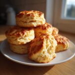 A stack of golden-brown, fluffy tea biscuits with visible layers, sitting on a white plate in natural light.