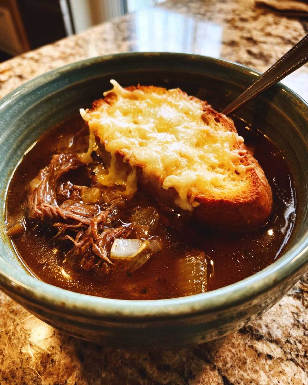 Close-up of a bowl of rich french onion beef stew topped with shredded beef, onions, and a slice of melted, cheesy toast.