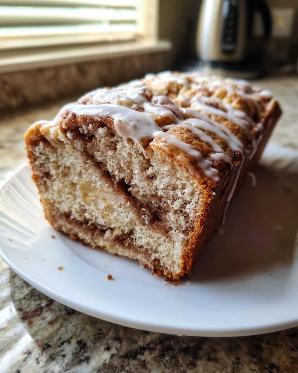 A freshly baked loaf of apple fritter bread with a cinnamon swirl and white glaze drizzle, sitting on a white plate.