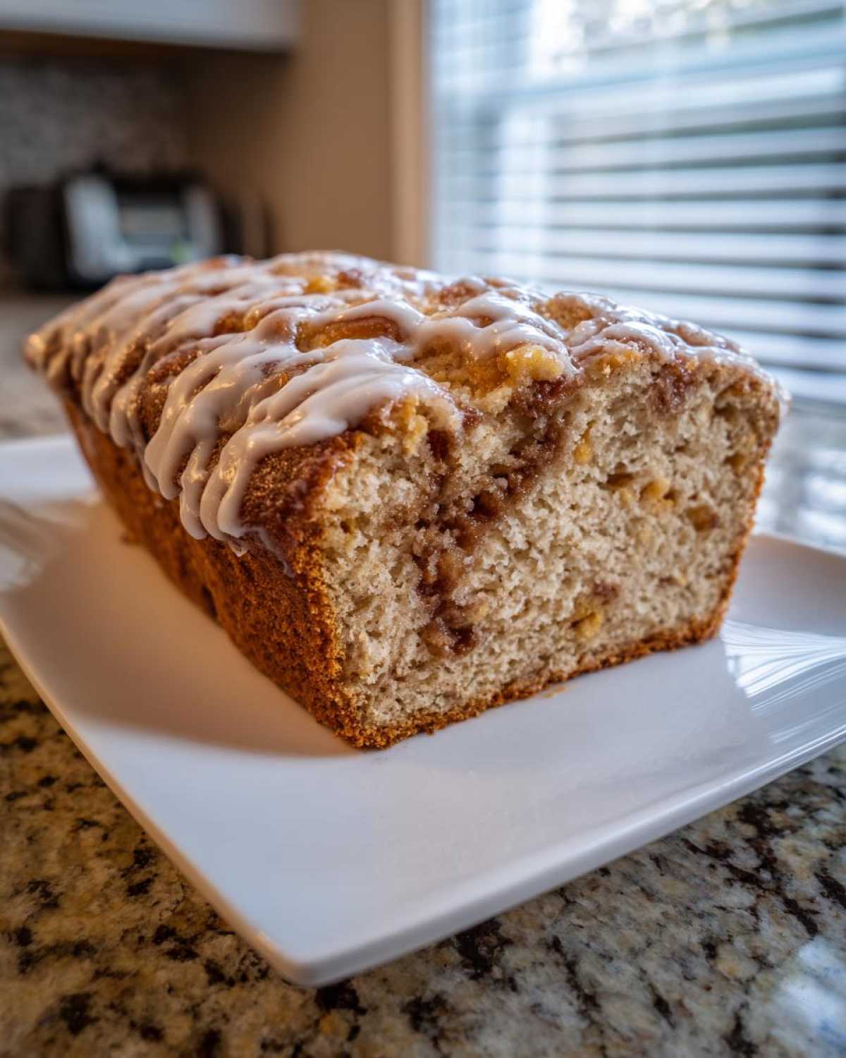 A freshly baked loaf of apple fritter bread with cinnamon swirl and white glaze, sitting on a white rectangular plate.