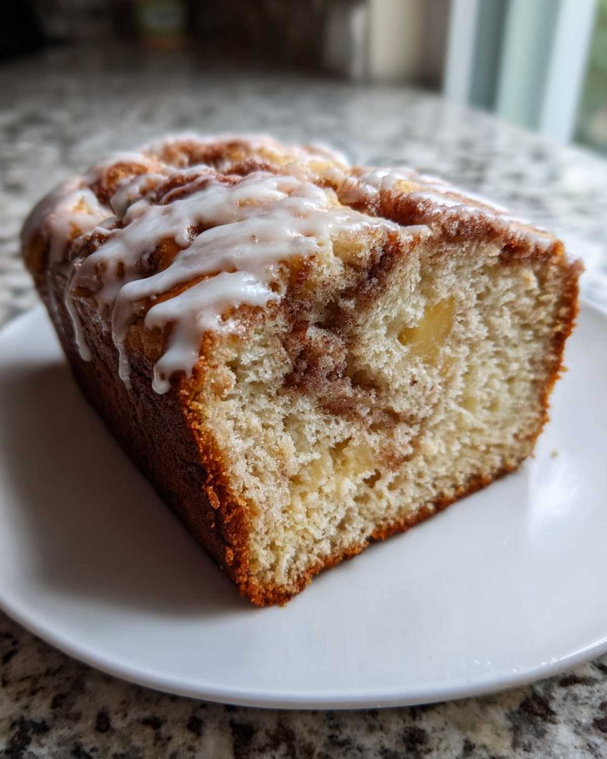 Close-up of a slice of moist apple fritter bread with cinnamon swirl and white glaze on a white plate.
