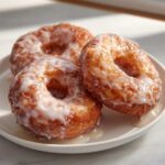 Three golden brown baked apple fritters generously coated in white glaze, resting on a white plate.