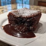 A square serving of rich chocolate cobbler oozing fudge sauce, dusted with powdered sugar.