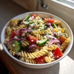 Close-up of a white bowl filled with colorful greek pasta salad featuring fusilli, tomatoes, cucumbers, red onion, olives, and feta.