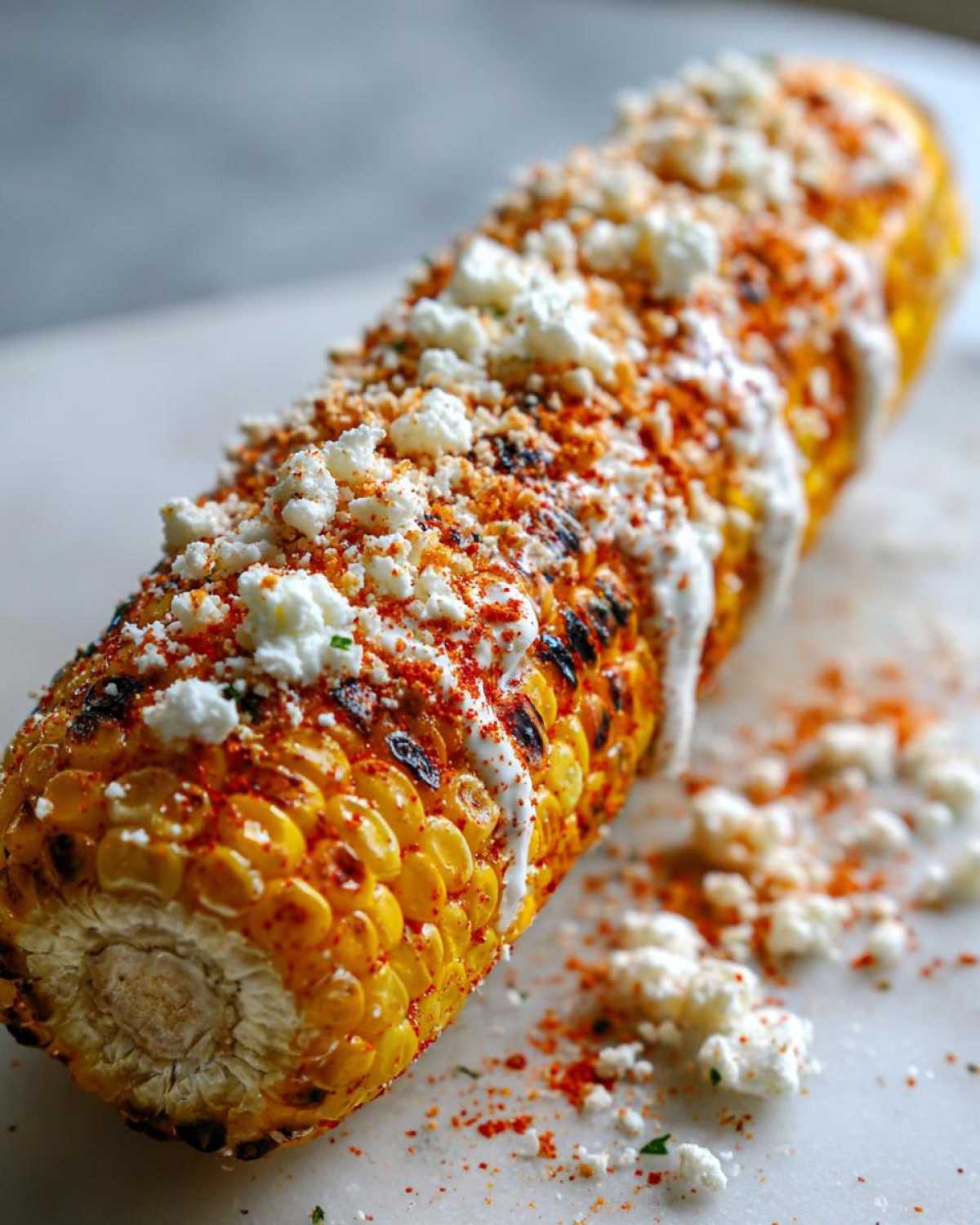 Close-up of a grilled ear of Mexican street corn heavily topped with white cheese, creamy sauce, and bright red chili powder.