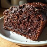 A moist, dark slice of Healthy Chocolate Zucchini Bread with visible chocolate chips on a light plate.