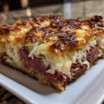 A thick, square slice of homemade Reuben bake featuring layers of corned beef, sauerkraut, and a deeply browned, cheesy topping.