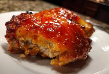A close-up of a piece of hot honey chicken with a thick, shiny, reddish-orange glaze on a white plate.