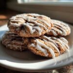 A stack of four freshly baked cinnamon roll cookies drizzled with white icing, sitting on a white plate.