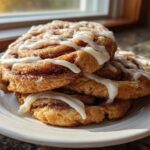 A stack of three freshly baked cinnamon roll cookies, generously drizzled with white icing, sitting on a white plate.