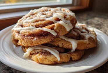 A stack of three freshly baked cinnamon roll cookies, generously drizzled with white icing, sitting on a white plate.
