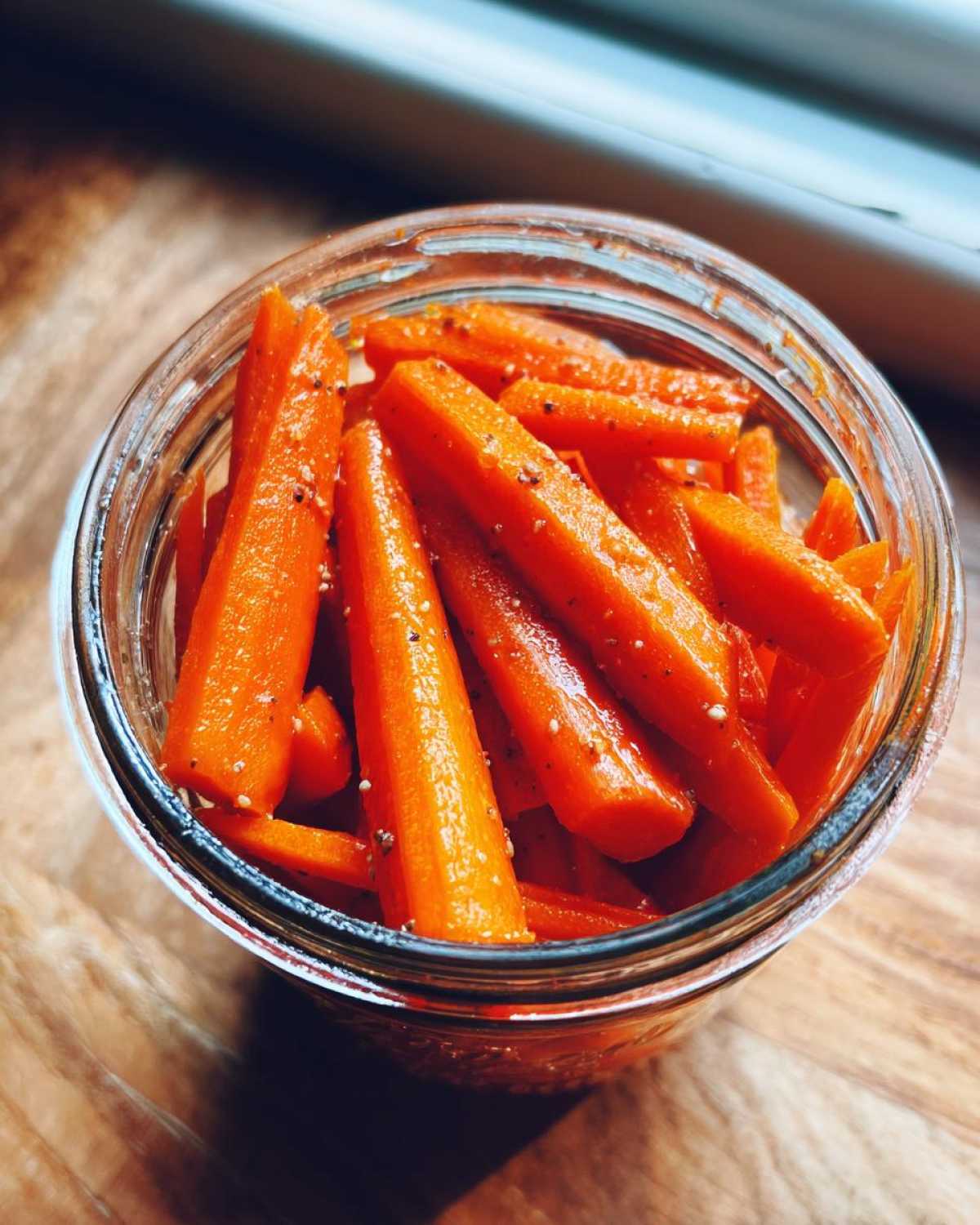 Overhead close-up of bright orange pickled carrots sticks seasoned with pepper inside a glass jar.