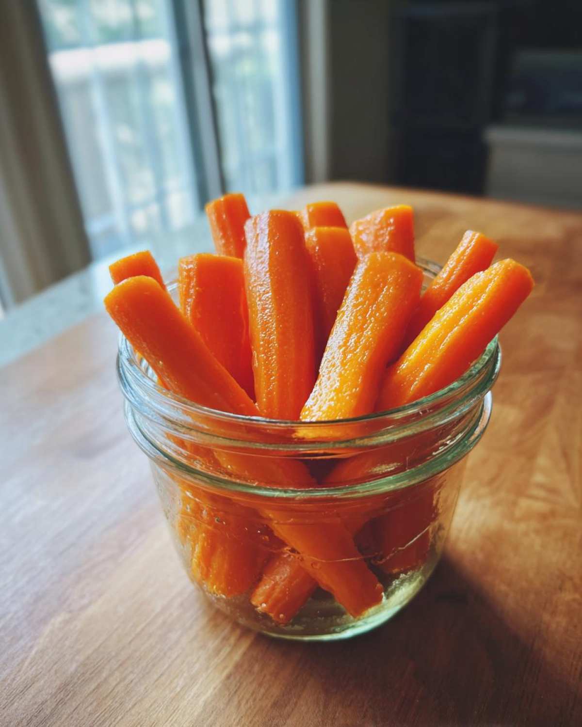 Close-up of bright orange pickled carrots sticks standing upright in a small glass jar on a wooden surface.