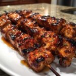 Close-up of juicy, glazed honey garlic chicken skewers resting on a white plate with extra sauce.