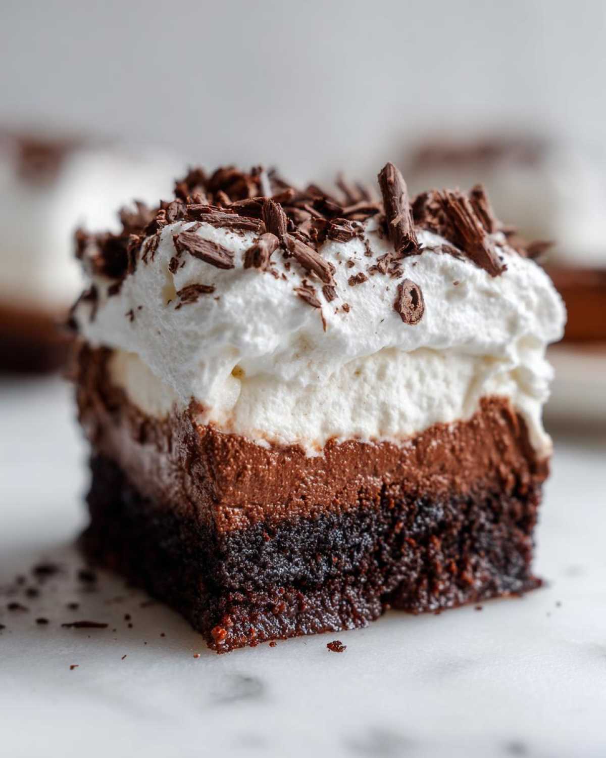 Close-up of a square slice of layered mud pie with a dark brownie base, chocolate mousse, whipped cream, and chocolate shavings.