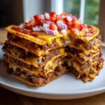 A tall, layered stack of mexican pizza showing melted cheese and ground beef filling, topped with fresh diced tomatoes and onions.
