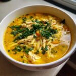 Close-up of a white bowl filled with vibrant yellow chicken orzo soup, topped generously with fresh parsley.