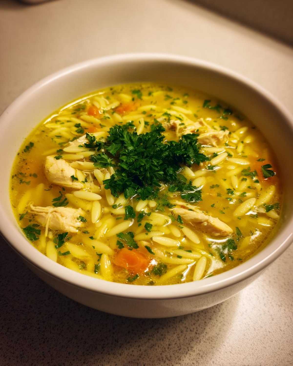 A close-up of a white bowl filled with bright yellow chicken orzo soup, featuring shredded chicken, carrots, and topped with fresh parsley.