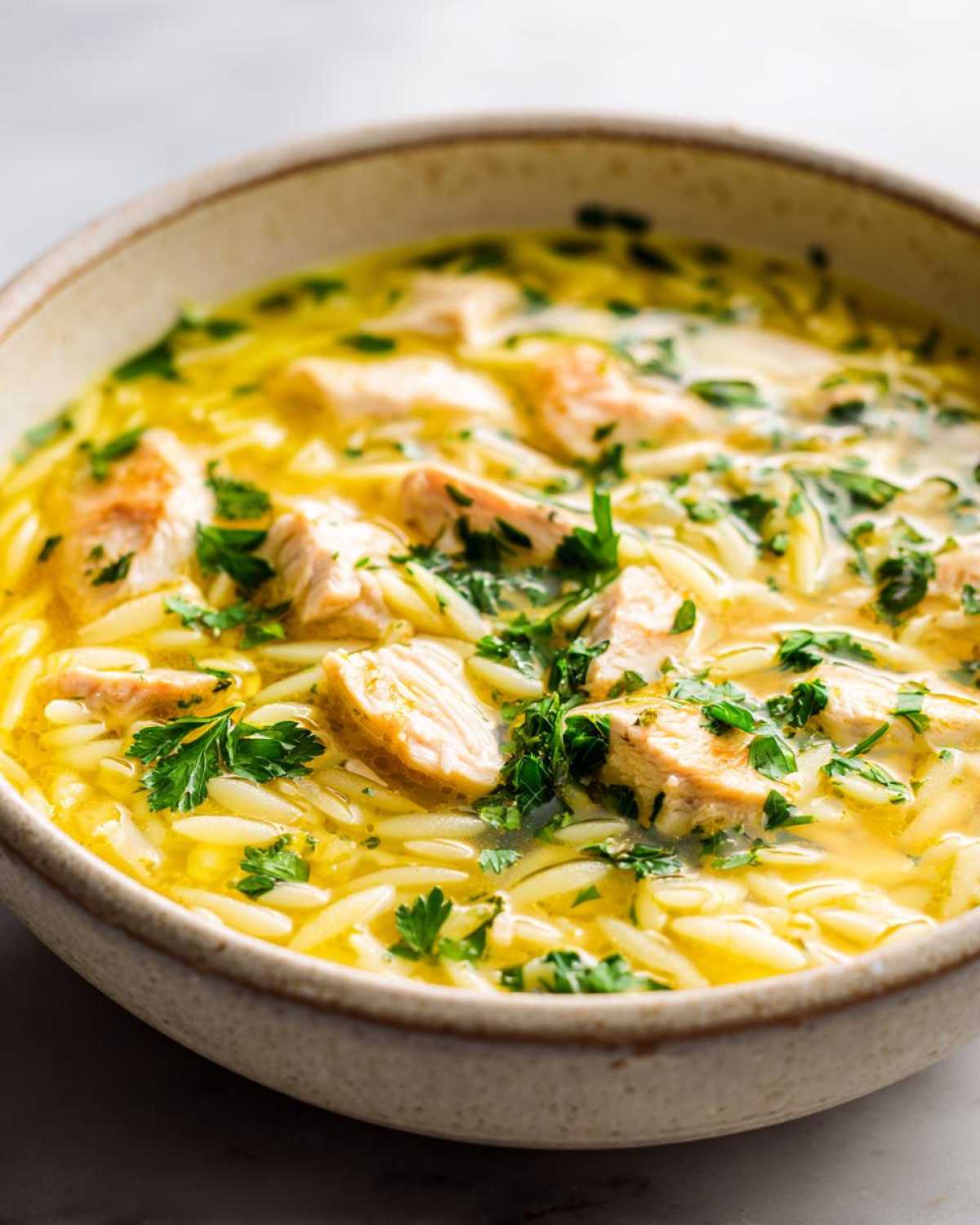 Close-up of a rustic bowl filled with bright yellow Lemon Chicken Orzo Soup, featuring chunks of chicken and fresh parsley.