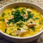 A close-up view of a bowl filled with bright yellow chicken orzo soup, featuring chunks of chicken and topped with fresh parsley.