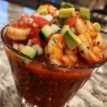 Close-up of a glass filled with vibrant Mexican shrimp cocktail, featuring plump shrimp, tomato sauce, avocado, and diced vegetables.