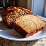 A close-up of a thick, moist slice cut from a golden brown banana bread recipe loaf on a white plate.