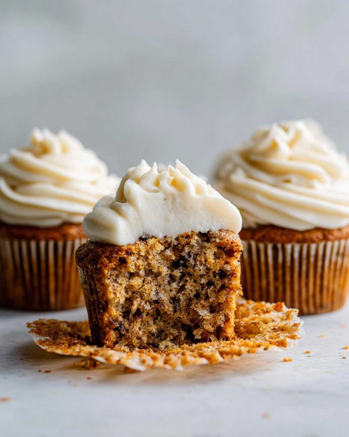 Close-up of a moist banana cupcakes cut in half showing the texture, topped with cream cheese frosting. Three banana cupcakes total.