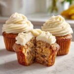 A moist banana cupcake cut in half showing the texture, topped with cream cheese frosting, with two whole banana cupcakes in the background.