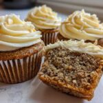 Close-up of moist banana cupcakes, one cut in half showing the texture, topped with cream cheese frosting.
