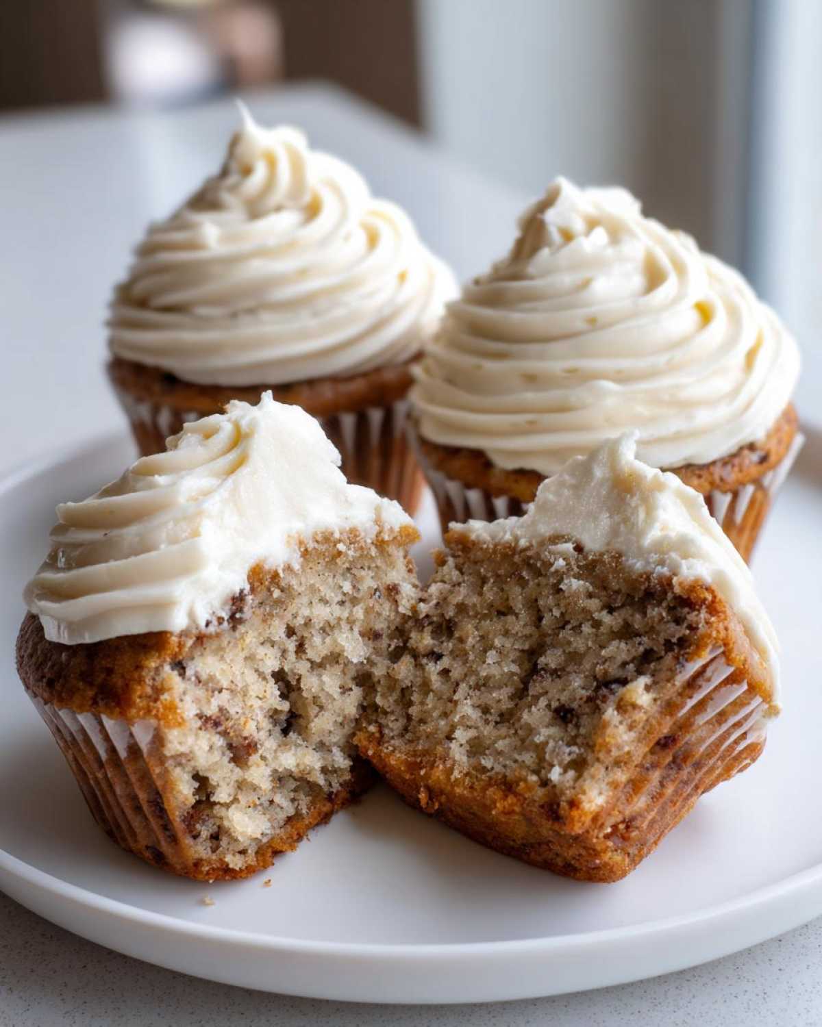Close-up of moist banana cupcakes, one cut in half showing the texture, topped with white frosting.