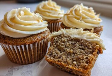 Close-up of moist banana cupcakes, one cut in half showing the texture, topped with cream cheese frosting.