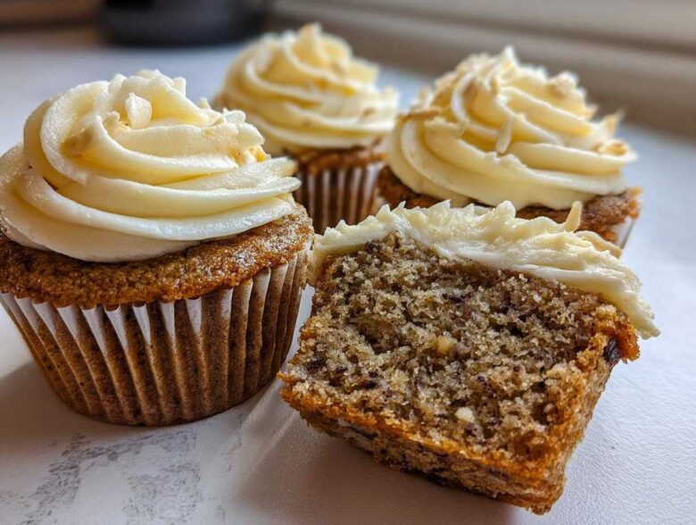 Close-up of moist banana cupcakes, one cut in half showing the texture, topped with cream cheese frosting.