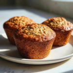 Three golden brown banana oatmeal muffins sitting closely together on a light-colored plate, highlighted by strong sunlight.