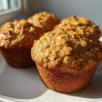 Four golden-brown banana oatmeal muffins with visible oat topping sitting on a white plate near a window.