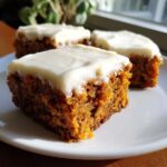 Close-up of three moist carrot cake bars topped with thick cream cheese frosting on a white plate.