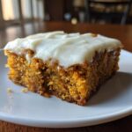 A close-up of a moist, square slice of carrot cake bars topped with thick cream cheese frosting on a white plate.