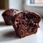 Close-up of a moist chocolate muffin cut in half, revealing chunks of melted chocolate chips inside the rich crumb of the chocolate muffins.